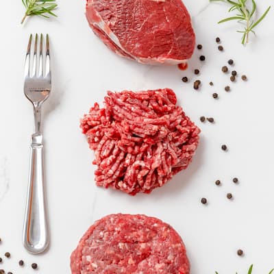 Vista cenital de un trozo de carne, un montoncillo de carne picada y un hamburguesa armada junto a un tenedor plateado y algunas especies esparcidas
